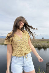 Woman stands in nature preserve, with wind blowing her hair in her face. She has a crochet lace top on, with a drawstring bottom, and pale denim shorts.  thumbnail