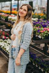 Woman in a white t-shirt and jeans leans against a shelf of flowers in a garden center. She wears a lacy crochet cardigan with elbow-length sleeves in a pale blue.  thumbnail