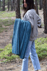 Woman standing in a cedar forest in a sweat shirt and jeans. She holds a crochet afghan draped over her left arm. The blanket is worked in a textured stitch in shades of blue.  thumbnail