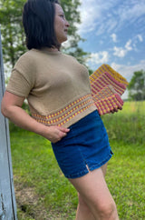 Woman in a tan knit t-shirt with colorful textured bands at the bottom leans against a building corner. She wears a denim mini skirt and holds a set of 4 device sleeves in matching colorwork. The trees and grass are bright spring green and the sky is blue with some wispy white clouds.  thumbnail