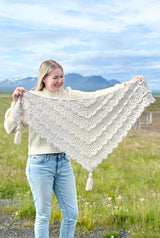 Woman in a pale sweater and jeans hold up an intricate lace crochet shawl, with tired ruffles. The shawl has tassels at each point. She stands in a green meadow with mountains in the background.  thumbnail