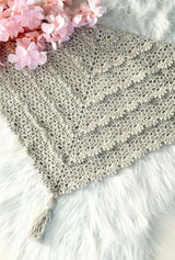Flat lay of romantic lace crochet shawl, with tiered scalloped ruffles and a tassel at the center point. It's laid on a white faux fur rug, with a bouquet of pale pink flowers in the upper left corner.  thumbnail