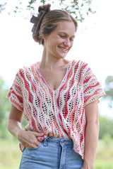 Woman standing outdoors with hair up in clip and wearing blue jeans. She has a lacy crochet top on in shades of cream, pink, and orange. thumbnail