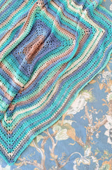 Multicolor crochet blanket lays on a floral patterned rug. The blanket is worked as a large rectangle from the middle out, in shades of rusty orange, blue, aqua, and a soft yellow.  thumbnail