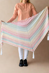 Model in front of a tan backdrop holds up a crochet shawl, with tassels on three points. The shawl is made in stripes of white and a self-striping yarn in a pastel rainbow.  thumbnail
