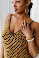 Close up of top half of crochet dress. The fabric is a granny cluster, with triangular front pieces and spaghetti straps. The model is wearing a cowrie shell bracelet and gold earrings.  thumbnail