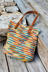 Multicolor crochet tote bag with faux leather handles sits on a weathered wooden deck, with some stones in the background. The bag is made in shades of yellow, orange, blues, and pale green.   thumbnail