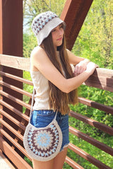 Woman in cream tank top and jean shorts leans on a wooden bridge railing. She wears a crochet bucket hat and a crossbody circular crochet bag in shades of cream, pale blue, and taupe.  thumbnail