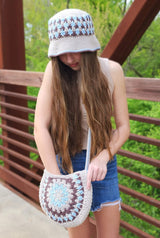 Woman standing on bridge with head angled down to show color stripes on hat bucket. Crown and brim are in cream. She wears a matching crochet crossbody bag with a cream body and side panels in colorwork to match hat.  thumbnail