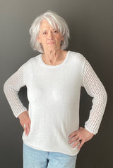 Woman in a white top and light wash jeans stands in front of a grey wall. The top is a knit long sleeve tee with a solid body and lace sleeves.  thumbnail