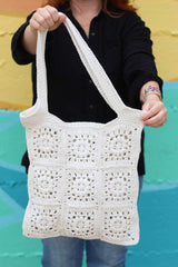 Close up of cream crochet bag made of crochet squares seamed together. A woman in a black top and jeans holds it toward the camera, standing in front of a painted wall.  thumbnail