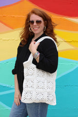 Woman in black top, jeans, and aviator sunglasses wears a cream crochet tote on her shoulder. She stands in front of a rainbow painted wall.  thumbnail