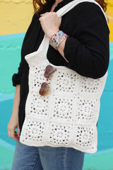 Close up of crochet shoulder bag, made of individual squares of cream yarn. Heart-shaped aviator sunglasses hang from the top of the bag.  thumbnail