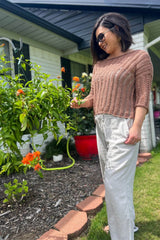 Woman stands near flower bed  in front yard. She wears a tan knit top and white flowy pants.  thumbnail