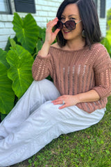 Woman in a tan lace knit top and white pants sits in a green yard in front of a large leafy green plant.  thumbnail