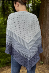 Back view of woman wearing large knit shawl. The top section is pale grey with an eyelet pattern, then there is a stripe section of pale and medium blue-grey. Then a section of medium blue-grey in a textured pattern, with a narrow stripe section of that color and dark blue-grey. The border is bark blue-grey with a cabled texture.  thumbnail