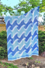 Crochet afghan with repeating wave motif hangs from a tree branch. The afghan is made in deep sky blue, robin's egg blue, and white.  thumbnail