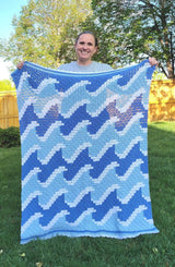 Woman holds crochet afghan toward camera, standing a yard of green grass.  thumbnail