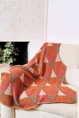 Crochet afghan draped over cream chair, in front of a white brick wall with a plant in a cold fireplace. The afghan has diamond motifs in rust, canyon red, beige, and taupe.  thumbnail