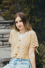 Woman in a pale yellow crochet lace cardigan, with short sleeves, sits on stone steps in front of a chain link fence.  thumbnail