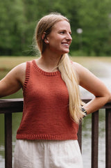 Woman in a rust tank top and cream pants stands in front of a wooden railing overlooking a body of water.  thumbnail