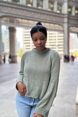 Woman in a pale green sweater and jeans stands with her hand in one pocket, in a city plaza.  thumbnail