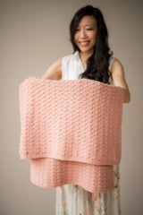 Woman in a white floral dress holds a pale pink baby blanket draped over her arms.  thumbnail