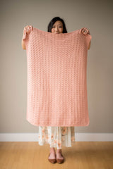 Woman holds baby blanket toward camera. The blanket is pink, and is made with a textured shell stitch. thumbnail