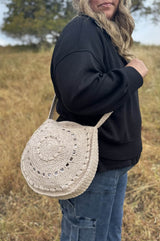 Woman in black sweatshirt and jeans wears a crochet bag crossbody style. The bag is pale beige, with a circular body and decorative texture.  thumbnail