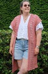 Front view of a sleeveless crochet cardigan, made in a peachy pink yarn with a lacy openwork pattern. It's worn over a white short sleeve top and jean shorts, by a woman standing in front of a green hedge.  thumbnail