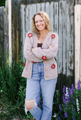 Woman standing in front of flower bed with worn wooden fence in the background. She wears a crochet cardigan over a brown tank top and jeans. The cardigan is pale grey with rust and pink flowers scattered on it.  thumbnail