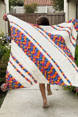Back view of young girl holding blanket behind her as she runs down sidewalk. The blanket is made in white and bright multi-color, with diagonal stripes of varying widths. Each corner has a large pompom.  thumbnail