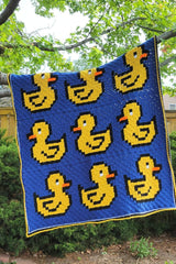 Crochet afghan hangs from tree branch in backyard. The afghan has a blue background with bright yellow rubber ducks, three rows of three, facing in alternate directions.  thumbnail