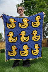 Woman holds crochet baby blanket in grassy yard. The afghan has a blue background, 6 yellow rubber ducks, and a striped border all around.  thumbnail