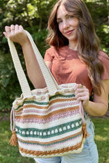 Woman in a rust colored t-shirt and jean shorts stands in a backyard. She holds a crochet tote with shoulder-length straps. The tote is ecru with stripes of amber, sage green, and pale pink. It cinches at the top with ties and tassels.  thumbnail