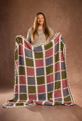 Woman standing in front of a warm tan studio backdrop holds a crochet afghan. The blanket has squares of medium grey-blue, olive green, and rose pink. Each block is bordered by white.  thumbnail