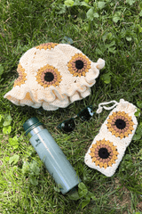 Flat lay of hat and pouch laying in grass, with a water bottle and sunglasses. The hat and pouch feature a sunflower motif on a cream background. The hat has a ruffled brim, and the pouch has a gathered top.  thumbnail