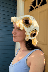 Woman in a blue tank top stands in front of a slate blue house with a mustard yellow door. She wears a crochet bucket hat with a ruffled brim, in cream. The hat body has sunflower granny squares in mustard, brown, and black.  thumbnail