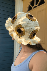 Back view of crochet sun hat showing sunflower granny square on top, and around the body of the hat. The flowers have a black center, then brown, then mustard yellow. The space between flowers is cream, and the brim is ruffled.  thumbnail