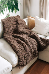 Chocolate brown afghan laid on cream couch, with white aad yellow throw pillows. The floor is a warm wood and there is a plant in the background.  thumbnail