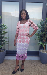 Woman stands on patio in front of glass doors with potted plants on either side. She wears high heels and a white slip dress, with a crochet lace dress over that. The dress is made in shades of pink, mauve, sky blue, and lilac purple.  thumbnail