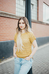 Woman leans against lamppost in front of brick building. She wears a crochet lace top in a sunny mustard yellow, with pale jeans.  thumbnail