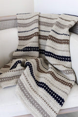 Striped crochet blanket draped over arm of chair. The blanket has a cream background with stripes of cool taupe, chocolate brown, and deep blue.  thumbnail
