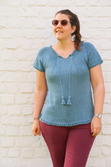 Woman with a ponytail and sunglasses stands in front of a white painted brick wall. She wears a blue knit top and burgundy pants.  thumbnail