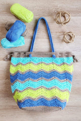 Crochet bag laid on a wooden table top, with decorative rope balls and four skeins of yarn above, on either side of the handles. The body of the bag has a ripple pattern with stripes of taupe, lime green, aqua, and deep sky blue.  thumbnail