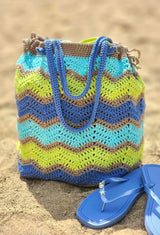 Crochet ripple bag sitting on beach, with a pair of blue flip flops. The bag has a drawstring top and short handles.  thumbnail