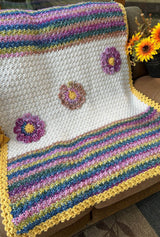 Crochet blanket laid over armchair indoors, with a bouquet of sunflowers on a side table. The blanket has a cream middle with three flowers applied in a diagonal line. The top and bottom of the blanket are stripes of pink, blue, green, and purple. The entire blanket has a yellow border.  thumbnail