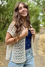 Woman in a blue top and denim jeans standing on a nature trail. She wears a lacy crochet sleeveless cardigan in cream.  thumbnail