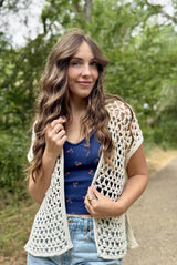 Woman wearing an open sleeveless crochet cardigan. It has a very open lace stitch and is made in a cream color.  thumbnail