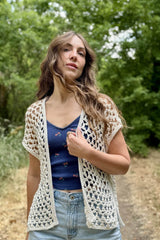 Woman in a blue top and jeans, with wind blowing her hair, standing on a dirt path through trees. She also wears a cream crochet sleeveless cardigan.  thumbnail